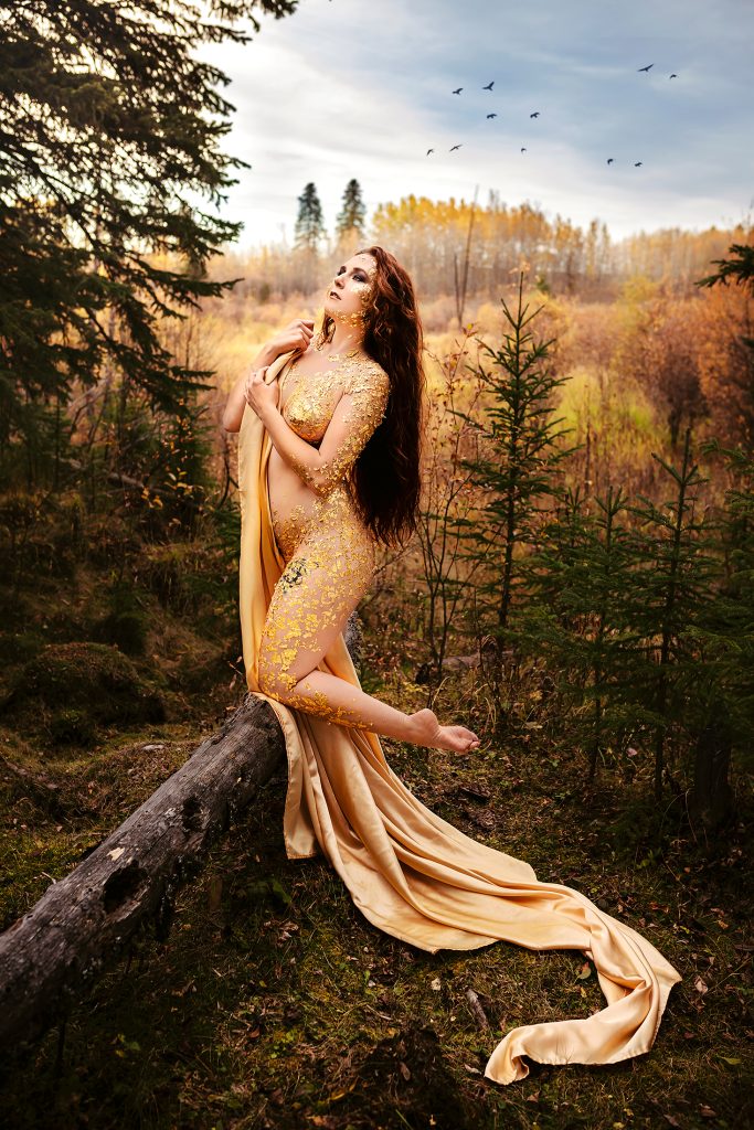 Woman with very long dark auburn hair wearing gold-leaf body art and draped fabric in an autumn forest.